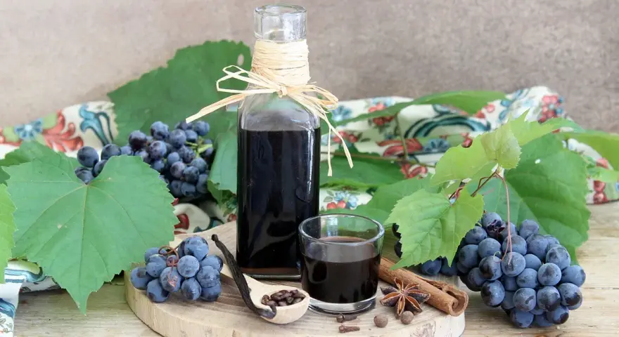 Mosto cotto ricetta: sciroppo scuro in bottiglia e bicchierino, con uva, spezie e cucchiaio di semi.