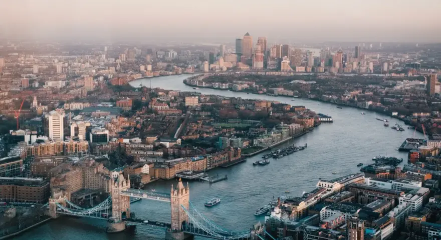Stunning drone photography and videography captures London's iconic Tower Bridge and the winding Thames River, with the city skyline stretching into the hazy distance.