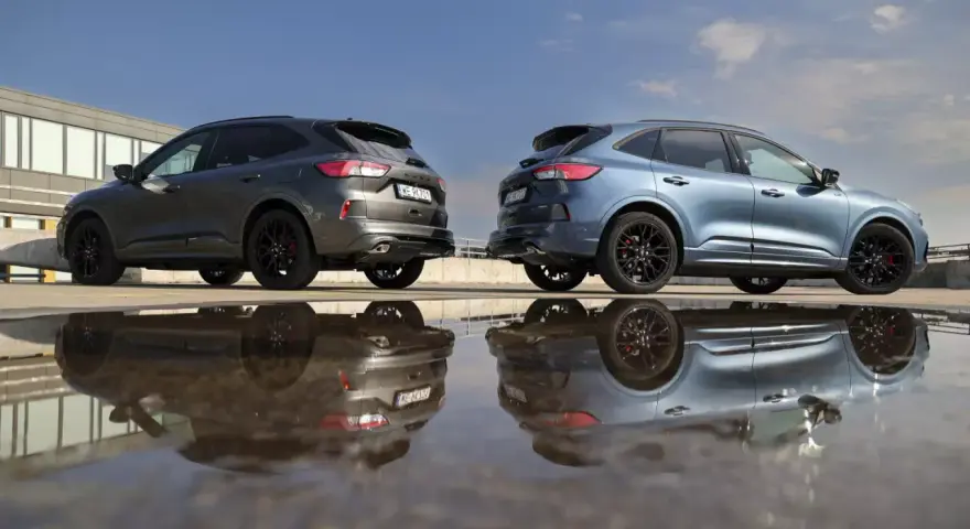 Dwa Fordy Kuga, jeden szary, drugi niebieski, stoją obok siebie na parkingu. Ich odbicia w kałuży tworzą surrealistyczny obraz. Oba modele to wersje hev.
