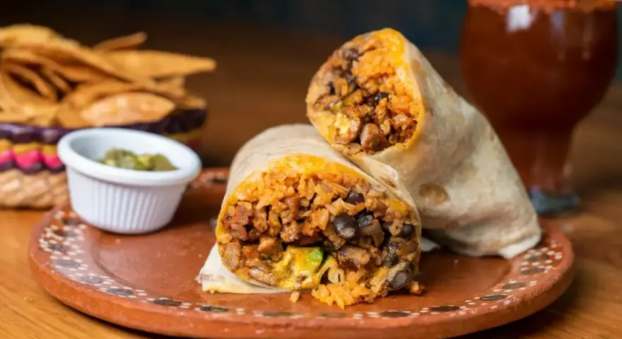 Pyszny burrito przepis: soczyste mięso, ryż, fasola i awokado zawinięte w tortillę. Obok chipsy i sos.