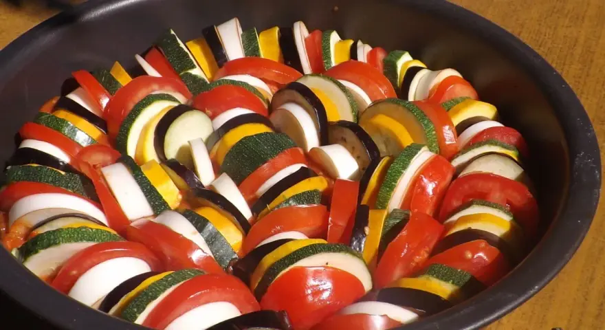 Un plat de légumes coupés en rondelles, prêt pour une recette de ratatouille digne du film. Tomates, courgettes, aubergines et oignons s'alternent.