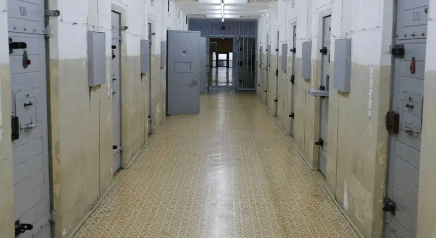 Korytarz więzienny z celami, gdzie może odbywać się tymczasowe aresztowanie. Widać drzwi z numerami, kraty i metalowe zabezpieczenia.