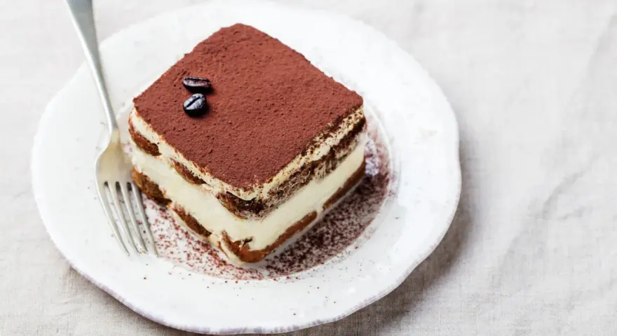 Una fetta di tiramisù, preparata con 500 g di mascarpone, guarnita con cacao e chicchi di caffè, su un piatto bianco con forchetta.