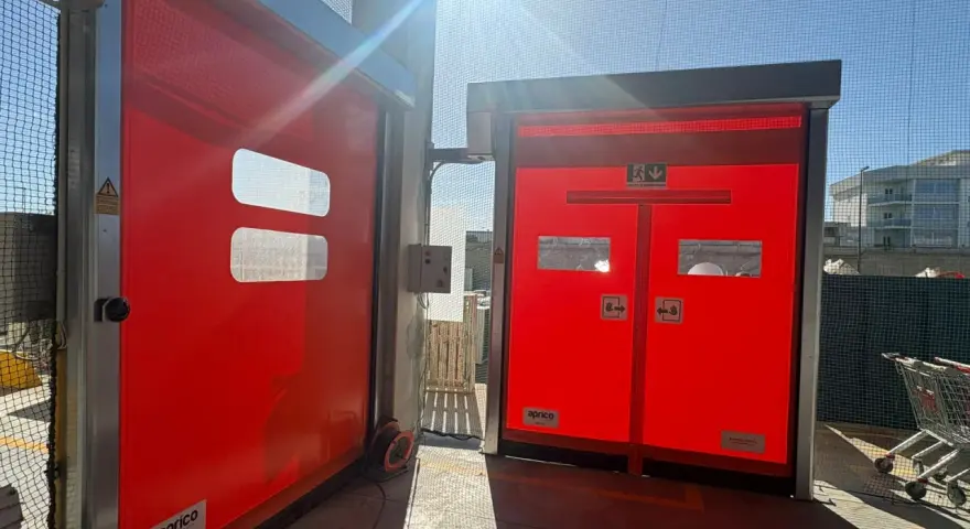 Due porte rosse automatiche, una aperta, mostrano come alzare una porta dal pavimento. Luce solare intensa.