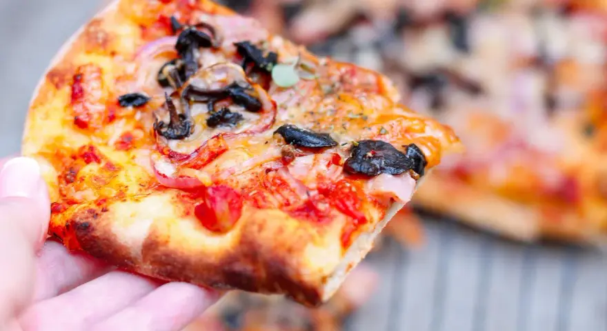 Kawałek pysznej pizzy funghi z pieczarkami, cebulą i pomidorami, trzymany w dłoni.