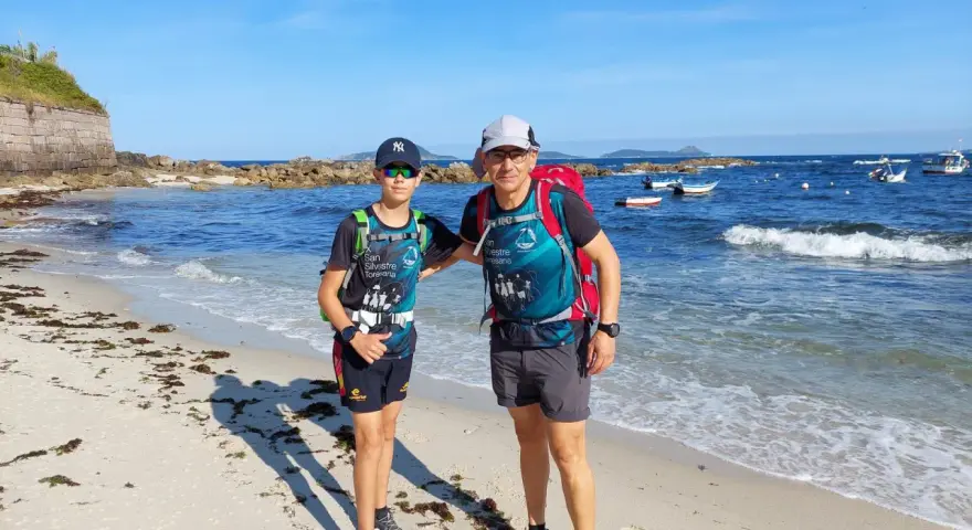 Dos excursionistas, padre e hijo, disfrutan de la playa en la etapa Baiona Vigo. El mar azul y el cielo despejado acompañan su aventura.