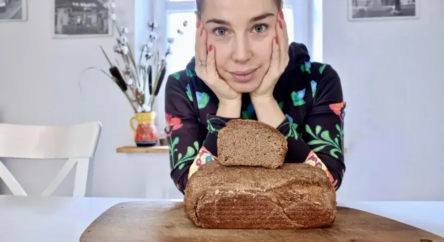Kobieta z dłońmi podpartymi pod brodą patrzy na pyszny chleb wieloziarnisty. Jeden bochenek leży na drugim, na drewnianej desce.
