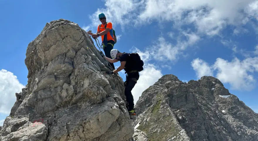 Zwei Bergsteiger meistern den **Hindelanger Klettersteig** auf felsigem Grat unter blauem Himmel mit Wolken.