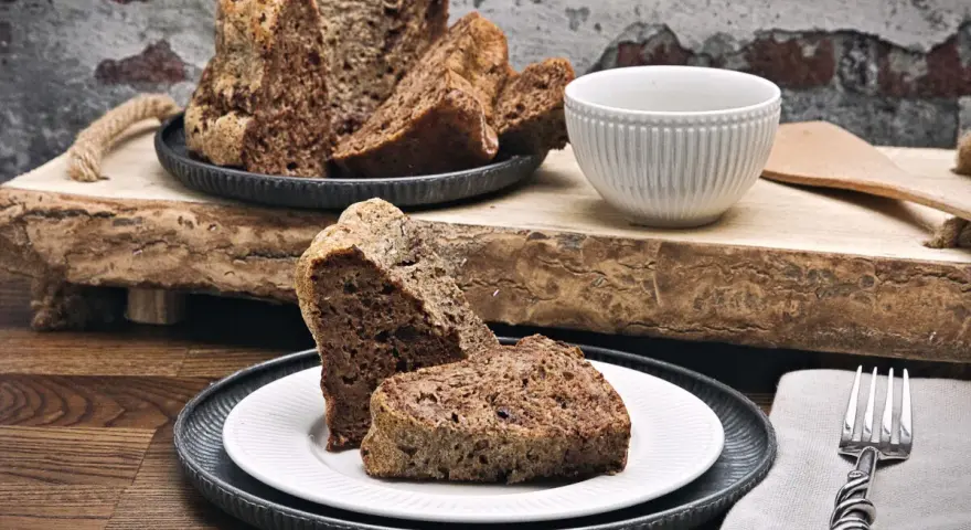 Zwei Stücke eines rustikalen, dunklen Diabetiker Kuchens auf einem Teller, daneben ein ganzer Kuchen mit angeschnittenen Scheiben.