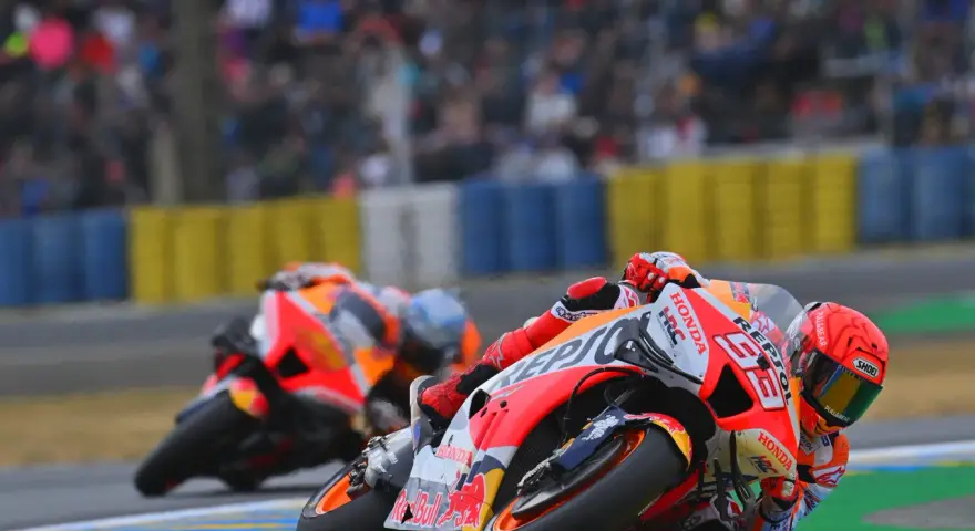 Dos pilotos de long lap motogp se inclinan en una curva, con el público borroso al fondo.