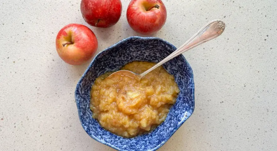 Compota de manzana casera en un cuenco azul, lista para disfrutar. Tres manzanas rojas frescas acompañan este delicioso postre.