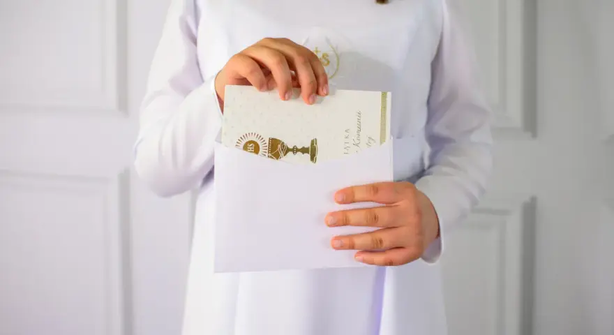 Dziecko w bieli wkłada kartkę z kielichem do koperty. To piękne krótkie życzenia na komunię, pełne symboliki.