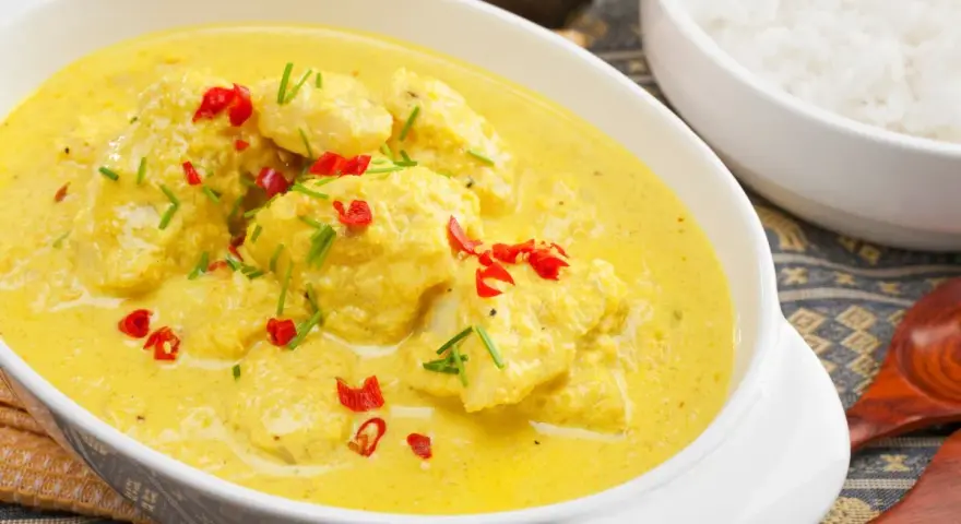 Delizioso pollo al curry con latte di cocco, guarnito con peperoncino rosso e erba cipollina. Accanto, una ciotola di riso bianco.