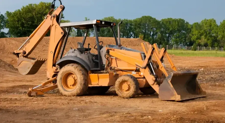 Pomarańczowa koparko-ładowarka na budowie. Czas pracy maszyny widać po zabrudzonych kołach i łyżkach.
