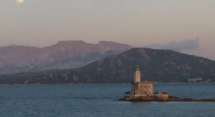 Faro solitario su un'isola sarda, con montagne sullo sfondo e luna piena. Perfetto per itinerari moto Sardegna 7 giorni.
