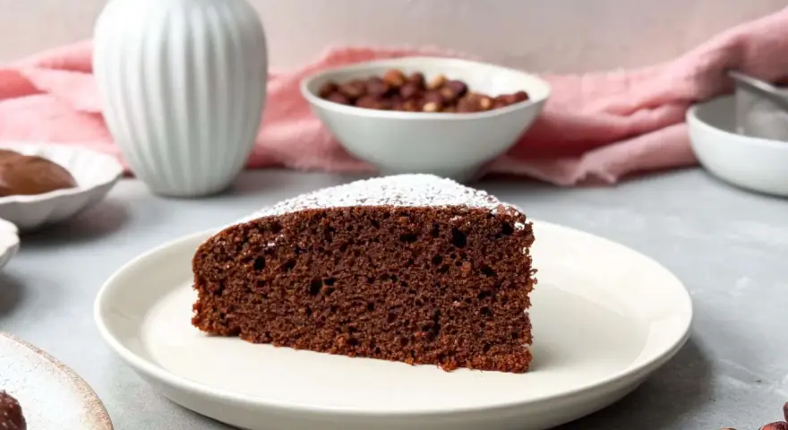 Ein saftiges Stück **Nutella Kuchen** mit Puderzucker bestäubt, daneben Nüsse und eine Vase.