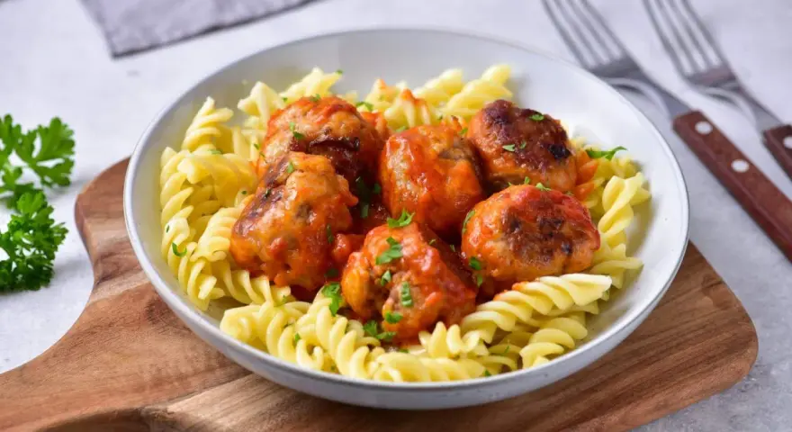 Pulpety w sosie pomidorowym jak u babci, podane z makaronem fusilli. Danie ozdobione posiekaną natką pietruszki.