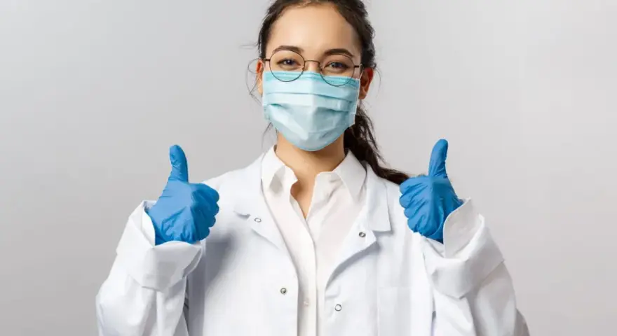 Uśmiechnięta pielęgniarka epidemiologiczna w masce i rękawiczkach pokazuje kciuki w górę.