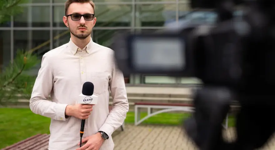 Młody dziennikarz w okularach trzyma mikrofon z logo "FLESZ" przed kamerą.
