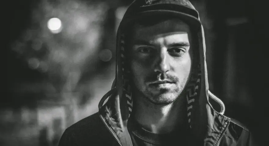 Mroczny portret mężczyzny w kapturze, z zacienioną twarzą i spojrzeniem, które zdaje się mówić o brudnych sercach i trudnych doświadczeniach.