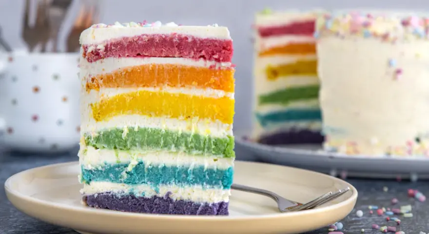 Ein Stück Regenbogen Kuchen mit bunten Schichten und Streuseln auf einem Teller.