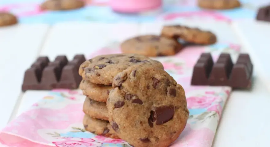 Una pila de galletas de chocolate caseras, con trozos de chocolate visibles, junto a barras de chocolate y un vaso de leche rosa.