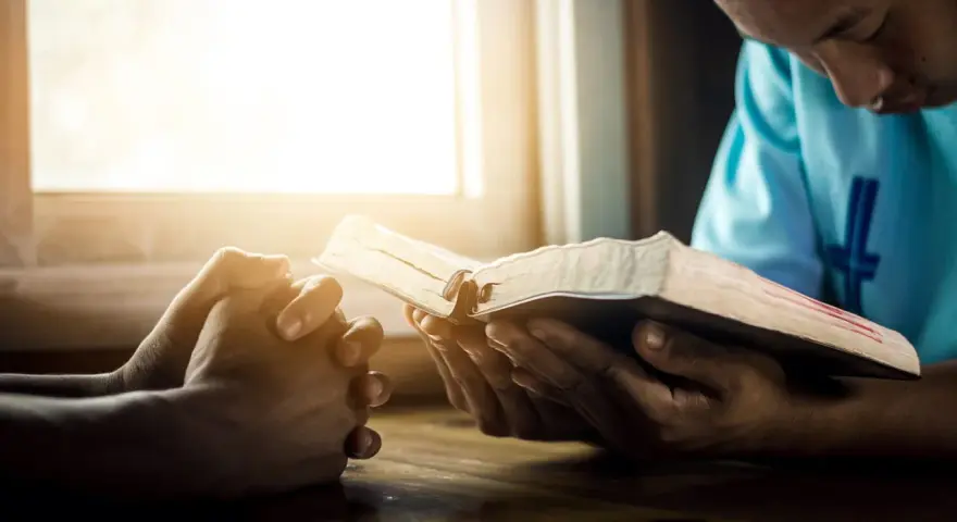 Hands clasped in prayer beside an open Bible, bathed in warm light. A moment of reflection, perhaps contemplating prayer names.