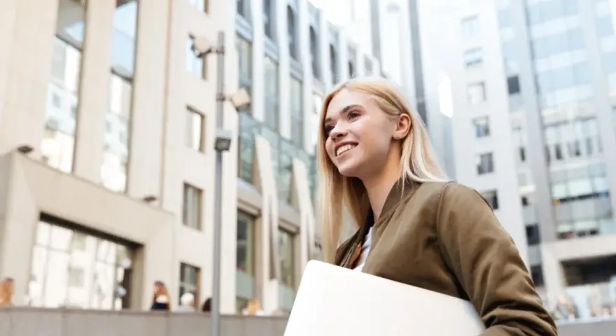 Giovane donna sorridente con laptop, cammina in una città moderna. Lavorare in Svizzera conviene, un futuro brillante ti aspetta.