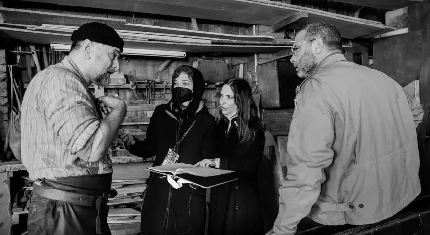 Foto di scena: un artigiano discute con due donne, una con mascherina, mentre un uomo osserva.