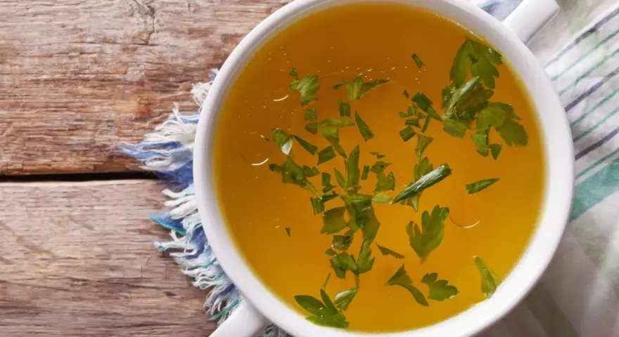 Aromaticzny bulion warzywny w białej miseczce, ozdobiony świeżą natką pietruszki, na drewnianym blacie.