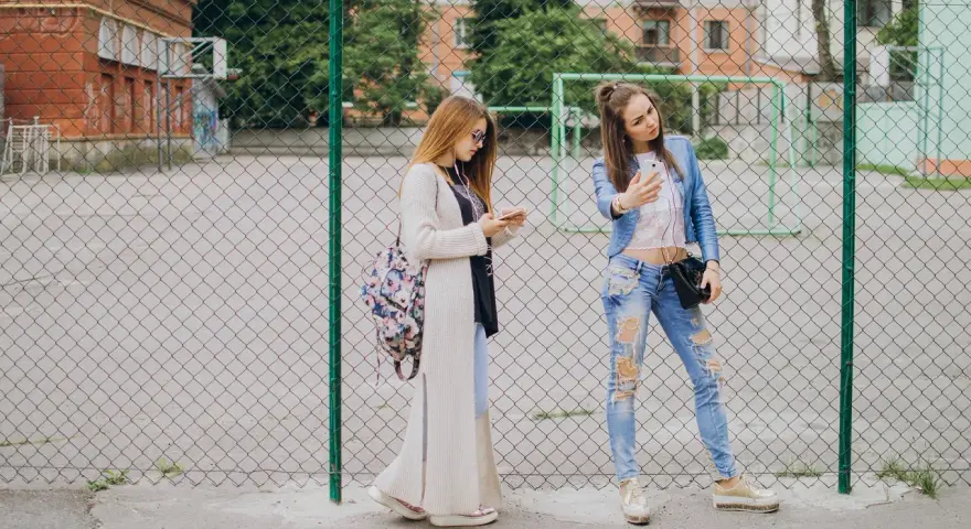Dwie dziewczyny w modnych stylizacjach, idealnych na outfit do szkoły, stoją przy płocie. Jedna robi sobie selfie, druga przegląda telefon.