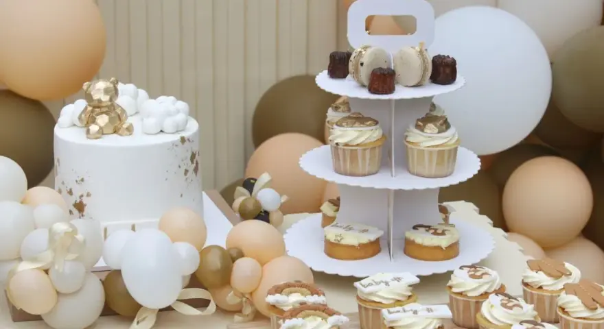 Biały tort z złotym misiem, babeczki i makaroniki na paterze, otoczone balonami w pastelowych kolorach.