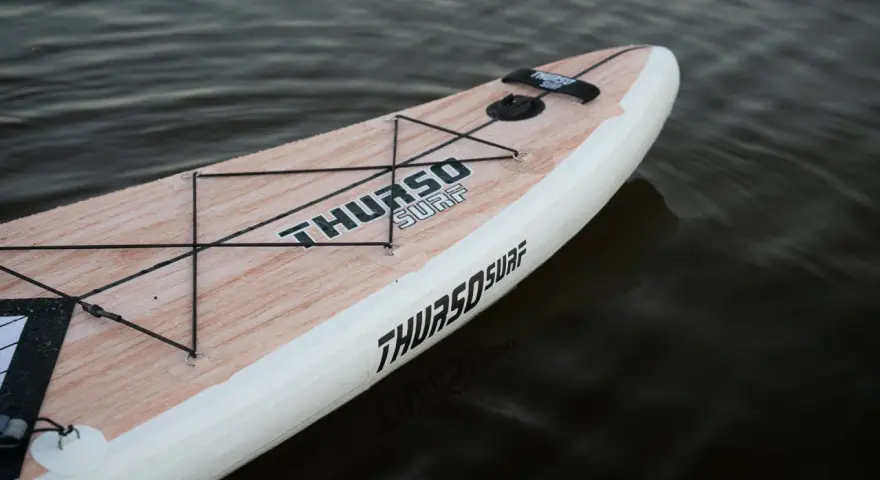 Ein aufblasbares SUP liegt im Wasser. Das Board hat eine Holzoptik und das Logo "THURSO SURF" ist gut sichtbar. Perfekt zum SUP lagern.