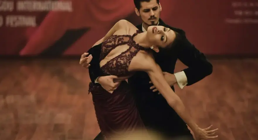 Para tańczy tango, jeden z rodzajów tańca towarzyskiego. Kobieta w bordowej sukni i mężczyzna w czarnym fraku wykonują skomplikowaną figurę.
