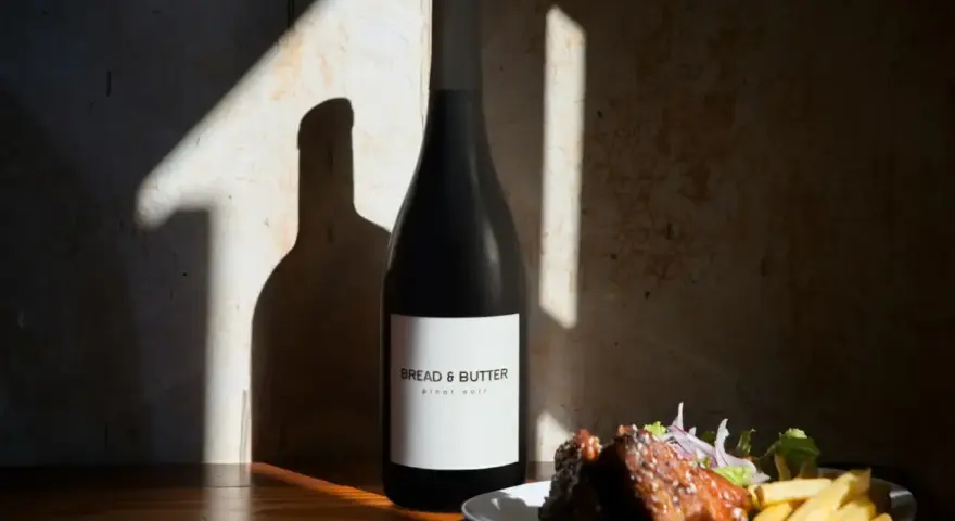 En flaska Bread & Butter Pinot Noir står bredvid en tallrik med revbensspjäll och pommes frites. Perfekt för en mysig kväll.