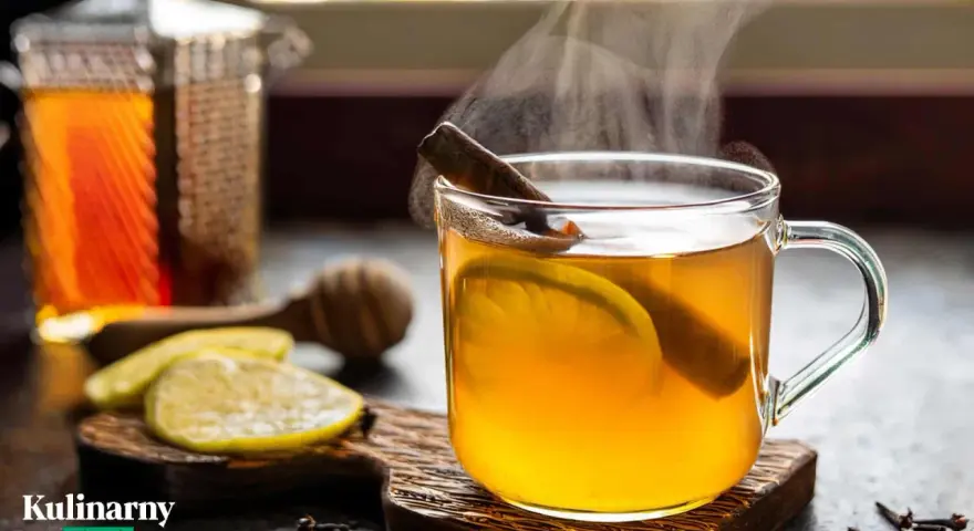 Rozgrzewająca herbata z whisky, z plastrem cytryny i laską cynamonu, paruje w szklance. Obok leżą cytryny i przyprawy.