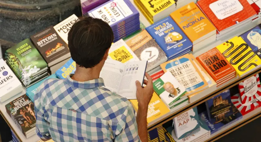 Hombre en una librería, rodeado de libros sobre finanzas y negocios, buscando las mejores editoriales para publicar un libro.