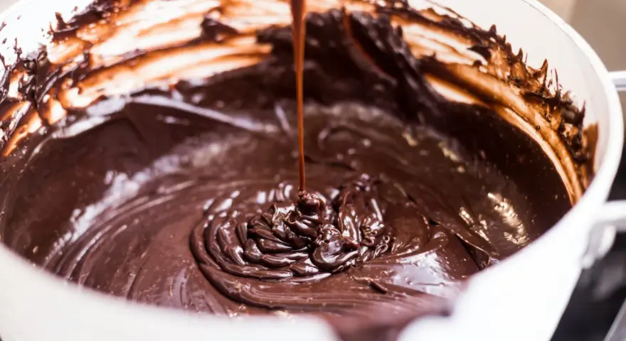 Gęsta, błyszcząca masa czekoladowa spływa z łyżki do białej miski. Idealna do fondant czekoladowy przepis.