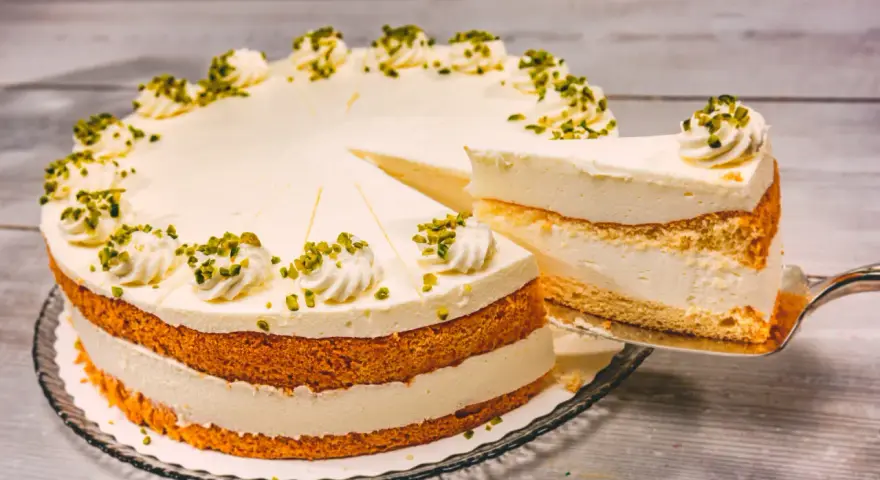 Eine Buttermilch Zitronen Torte mit cremiger Füllung und Pistazien-Deko. Ein Stück wird gerade herausgeschnitten.
