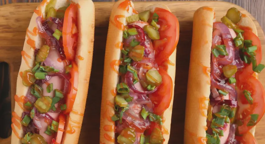 Trzy apetyczne hot dogi z frytkownicy powietrznej, z pomidorami, ogórkami, cebulą i szczypiorkiem, polane sosem.