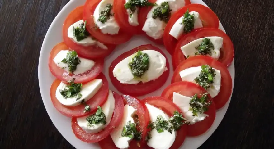 Pyszne pomidory z mozzarellą, ułożone na talerzu, ozdobione zielonymi ziołami. Idealna przekąska na lato.
