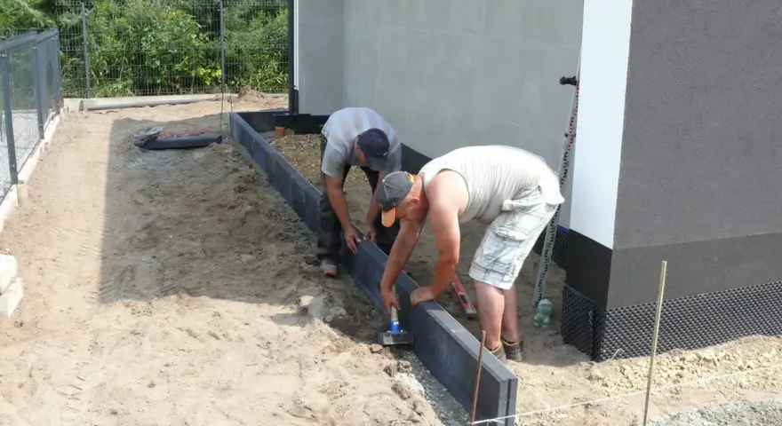 Dwóch mężczyzn układa obrzeża pod kostkę. Suchy beton pod kostkę jest przygotowywany do dalszych prac.