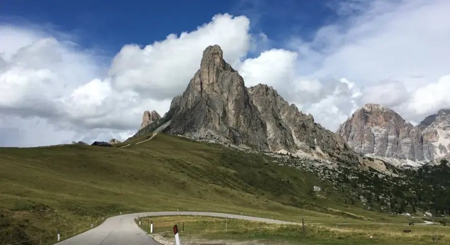 Strada tortuosa verso le Dolomiti, perfetta per una gita in moto. Montagne imponenti e cielo azzurro con nuvole bianche.