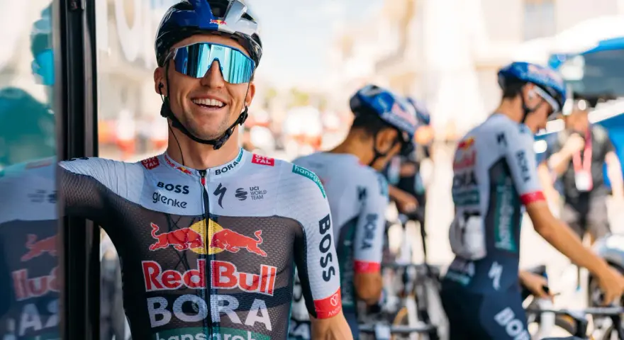 Cyclist smiles on a vuelta rest day, wearing a BORA-hansgrohe kit with Red Bull logos. Other team members and bikes are visible.