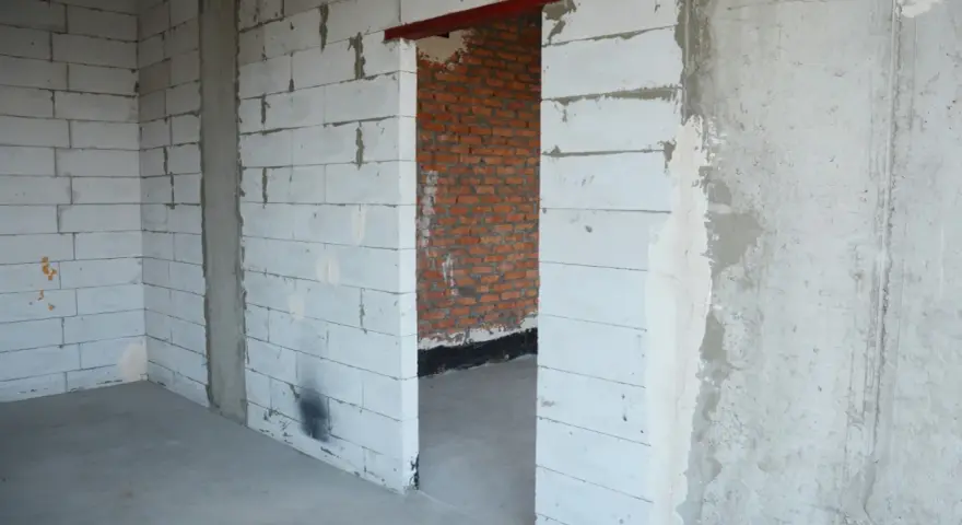 Wnętrze w budowie. Widoczny otwór drzwiowy z czerwonym nadprożem stalowym, ściany z białych pustaków i cegły.