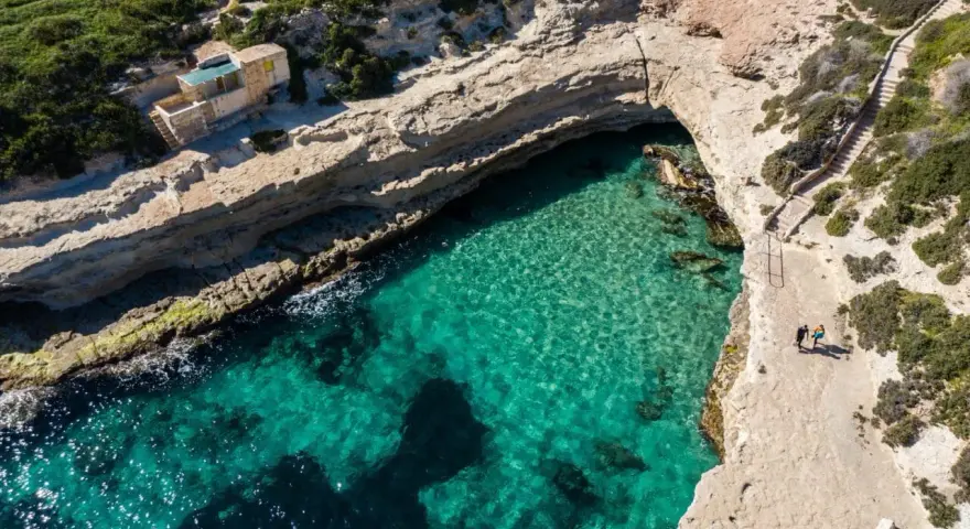 Malta plaże: turkusowa woda w zatoce, skaliste wybrzeże i schody prowadzące do ukrytej plaży. Dwie osoby spacerują po klifie.