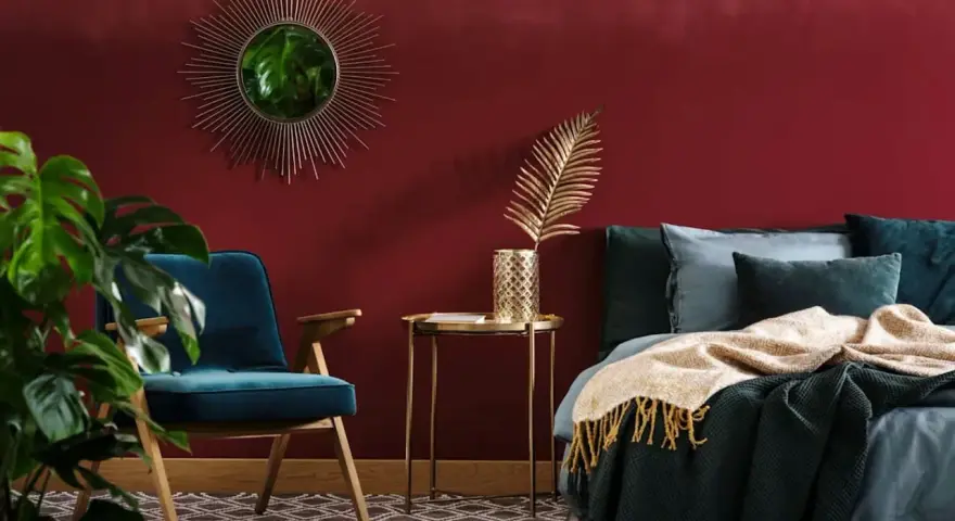 Habitación con pared de pintura a la tiza roja, sillón azul, cama con mantas y cojines, espejo sol y planta.
