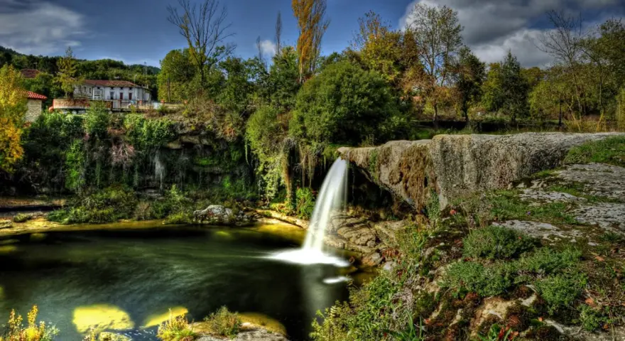 Cascadas en España: una casa rural se asoma entre la vegetación exuberante junto a una cascada que cae en un estanque de aguas verdes.