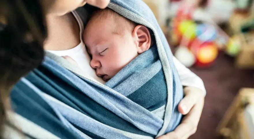 Ein Baby schläft friedlich in einem blauen Tragetuch. Der sanfte Tagesrhythmus des Babys wird durch die Nähe zur Mutter unterstützt.