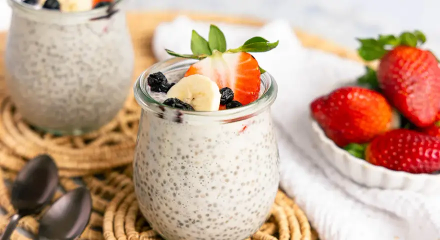 Zwei Gläser mit cremigem Chia Pudding, garniert mit Erdbeeren, Bananen und Blaubeeren. Daneben eine Schale frischer Erdbeeren.